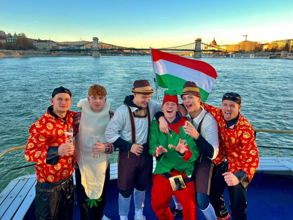 Five people toasting with beer glasses on a boat at sunset, enjoying a festive atmosphere during a stag boat cruise, during a budapest stag do.