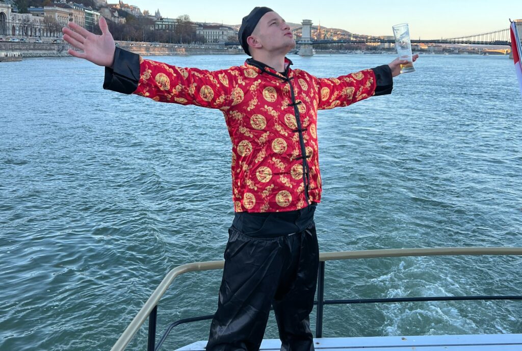 A man in a vibrant red patterned outfit poses with outstretched arms and a beer glass on a boat, enjoying the scenic Danube River and city views.