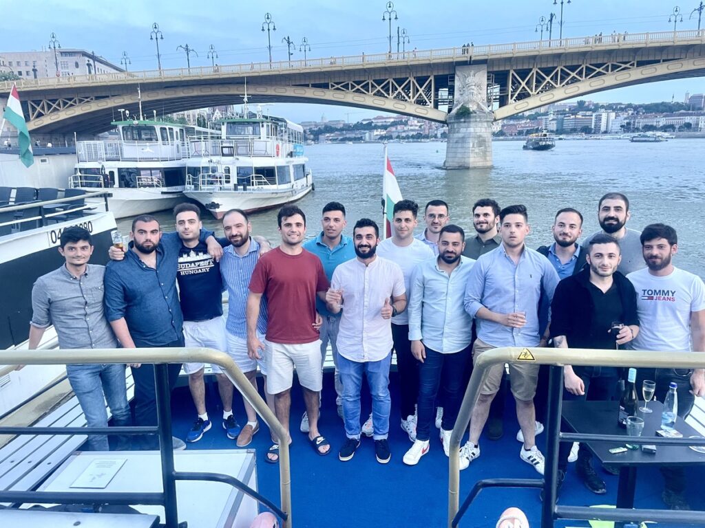 A group of friends posing for a photo on a boat during a Budapest stag do boat cruise, with the Margaret Bridge and the Danube River in the background.