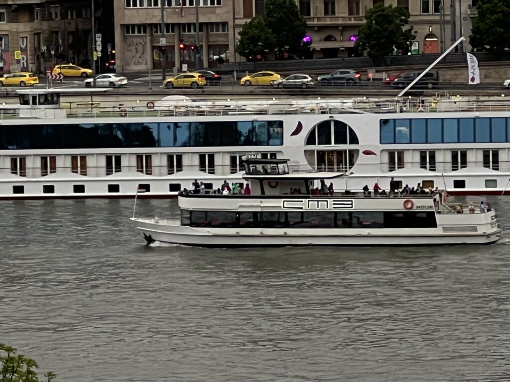 A passenger boat cruising along the Danube River in Budapest, with a larger river cruise ship docked in the background, and cars passing on the riverside street.