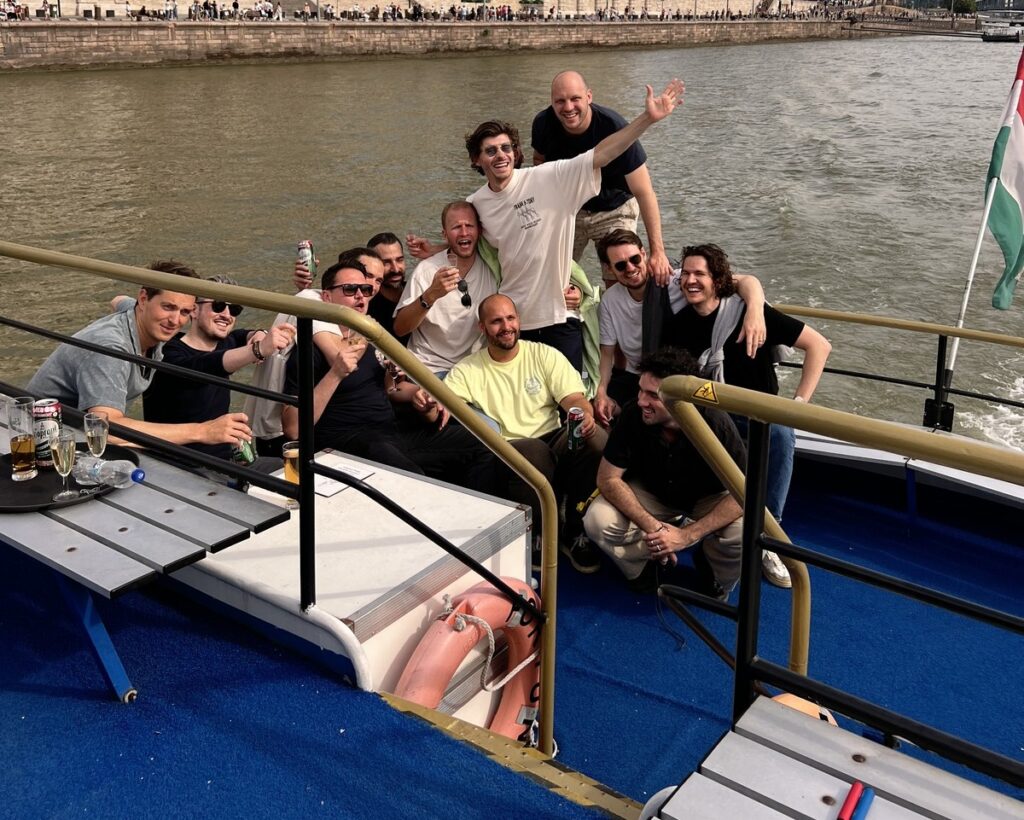 A lively group of friends enjoying drinks and celebrating on the deck of a boat during a Budapest stag do boat cruise, with the Danube River and city shoreline in the background.