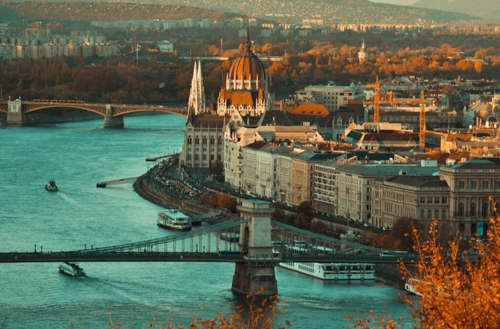 A picturesque aerial view of Budapest featuring the Hungarian Parliament Building, the Chain Bridge, and boats cruising on the Danube River during sunset.