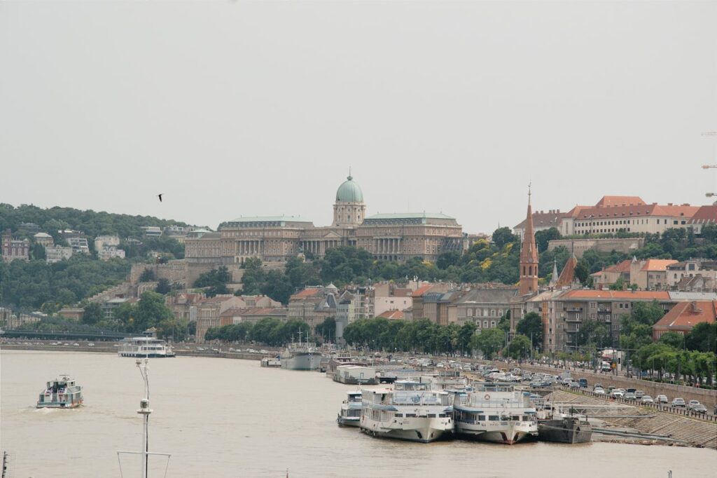 A stunning view of Buda Castle on a hill overlooking the Danube River, with moored boats and a riverside street, a must-see during a Budapest stag do boat cruise.